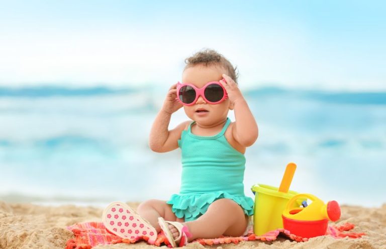 Toddler holding her sunglasses seated by the beach on a towel