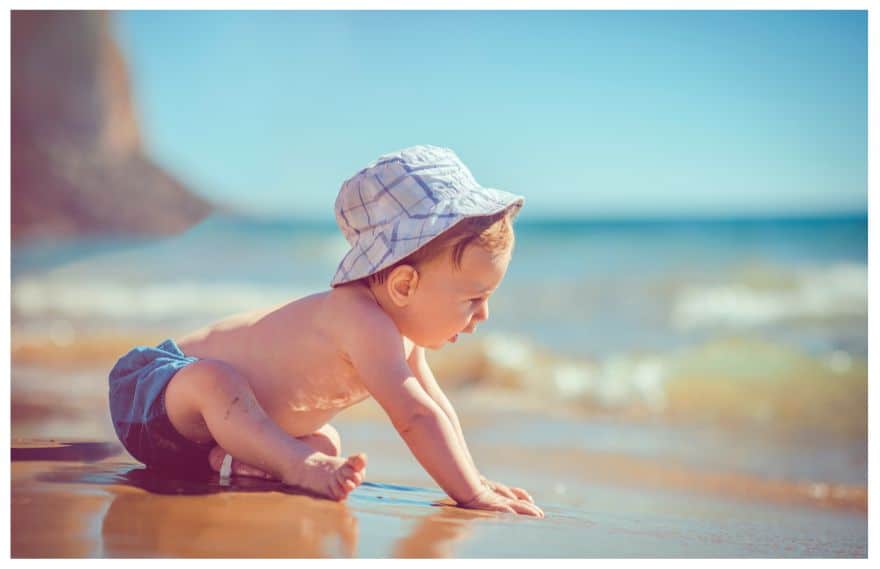 A baby boy sited at the beach with a hat and no shirt on